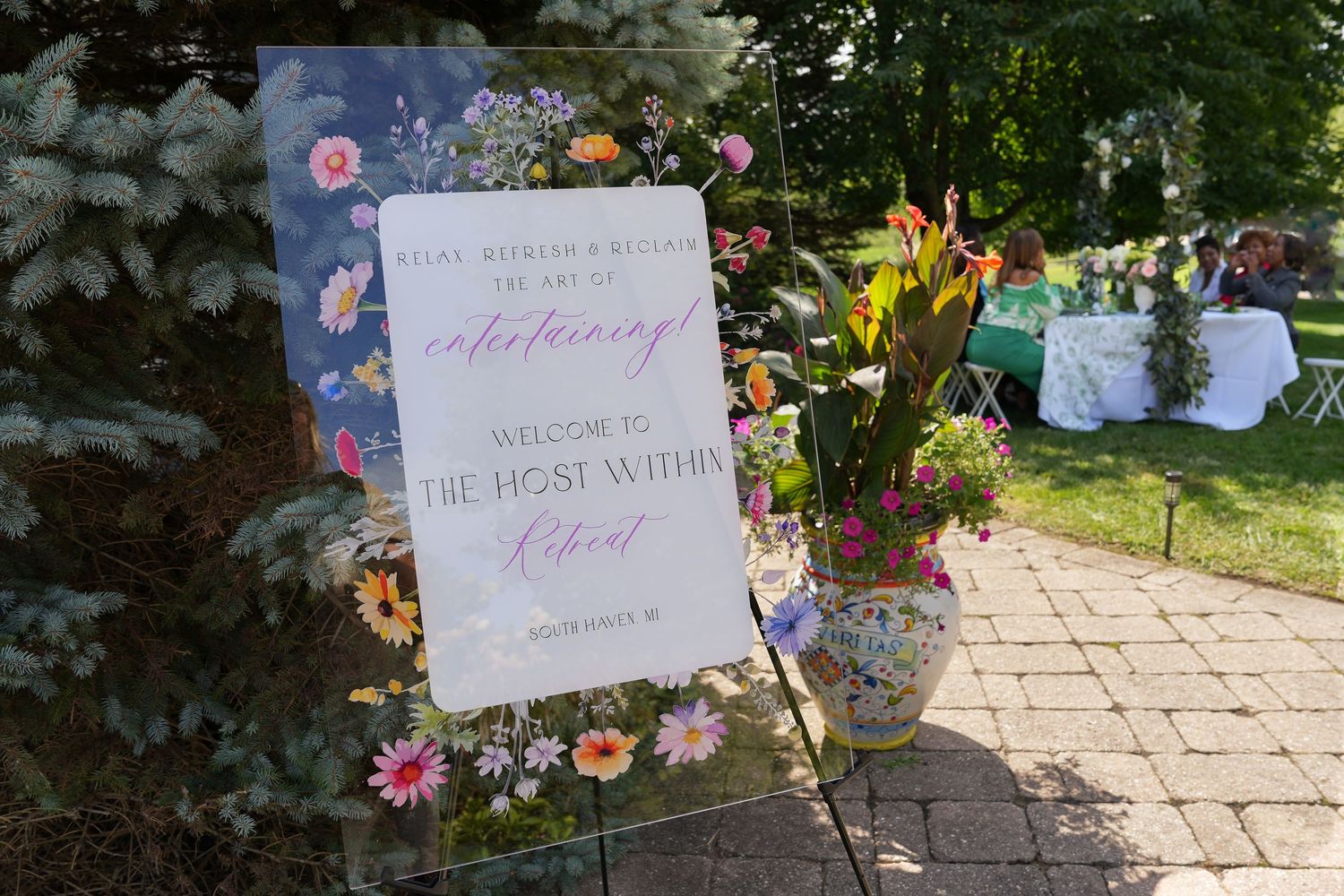 Floral welcome sign for The Host Within event on brick pathway with colorful flower arrangements outdoors.