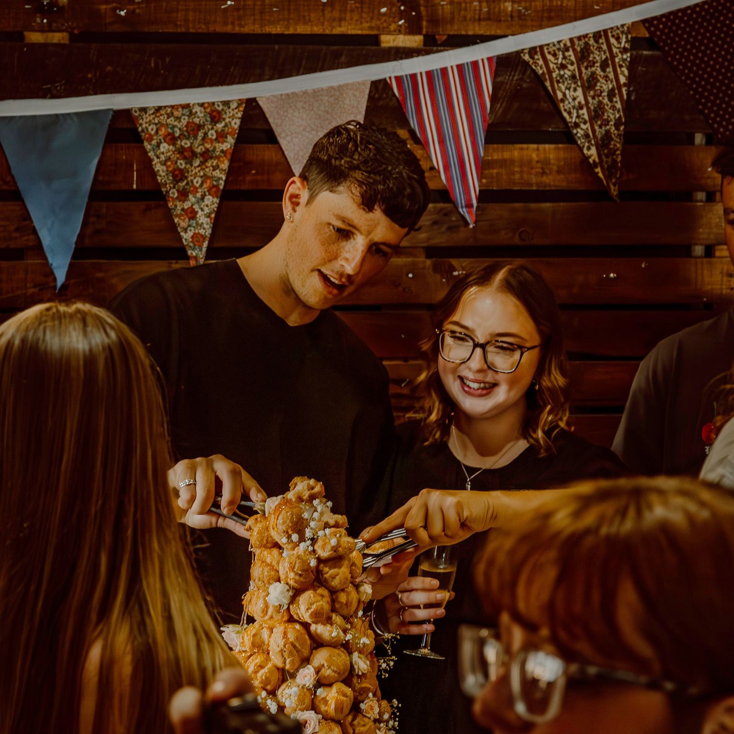 Friends gather around a croquembouche dessert tower under festive bunting decorations.