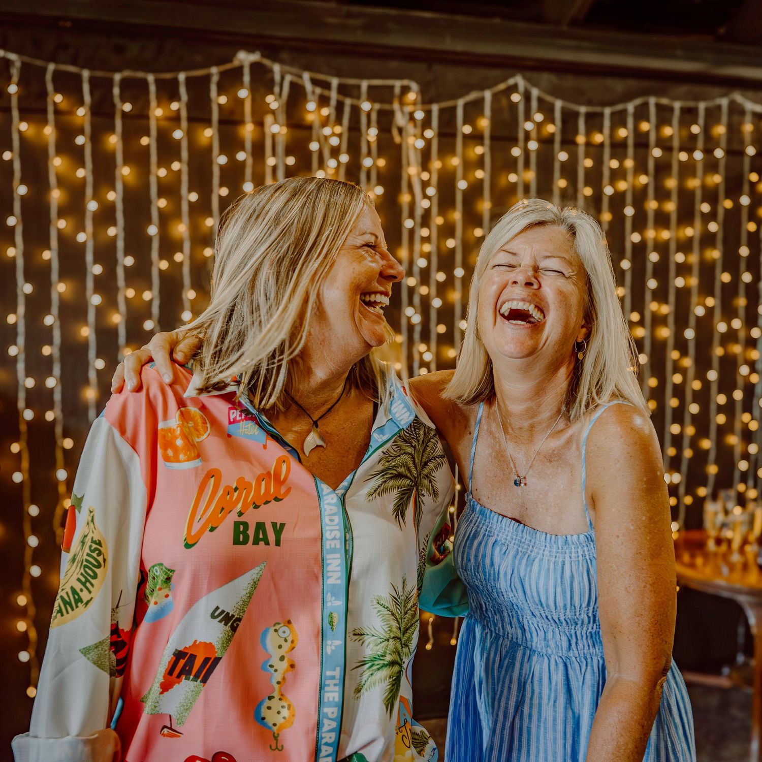 Two friends share a genuine laugh together in front of a wall of twinkling string lights.
