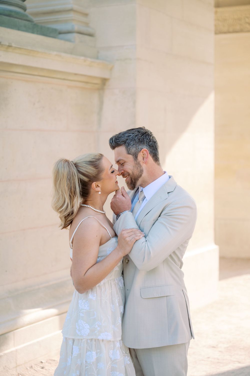 A couple shares a romantic moment near stone columns, the woman in a white dress and man in a beige suit almost kissing.