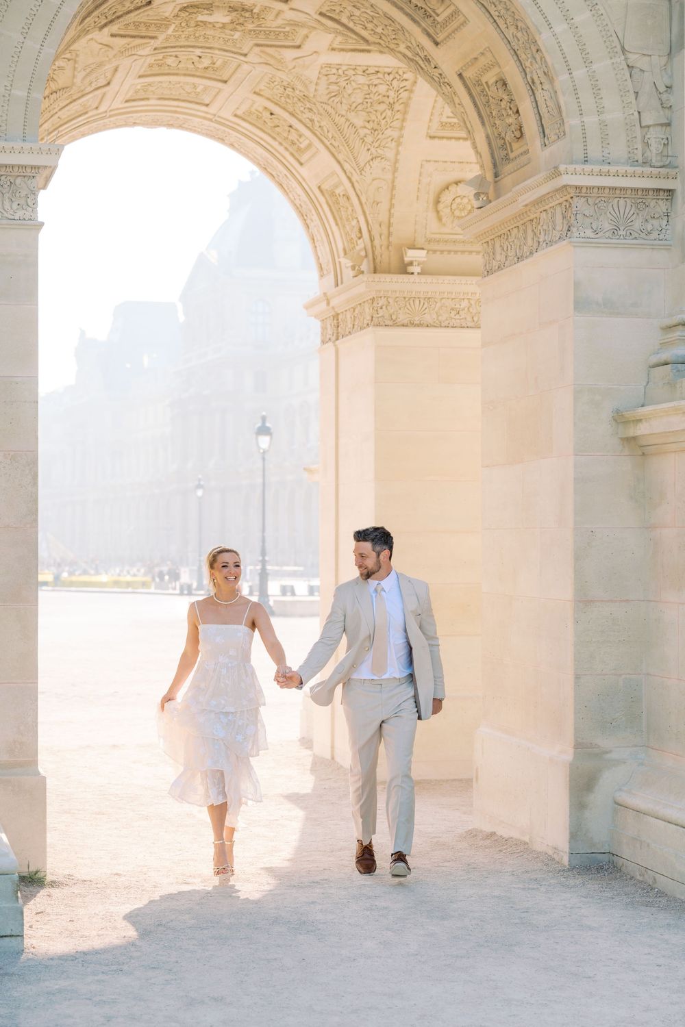 Couple holding hands walking under ornate stone arch at the Louvre in Paris, woman in white dress, man in light gray suit.