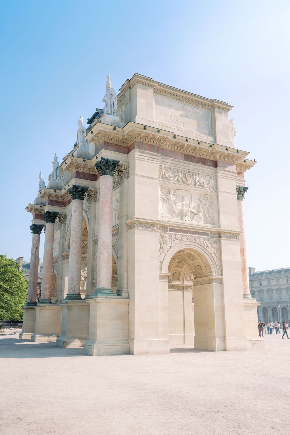 The Arc de Triomphe du Carrousel in Paris, a neoclassical triumphal arch with marble columns and relief sculptures under a clear blue sky.