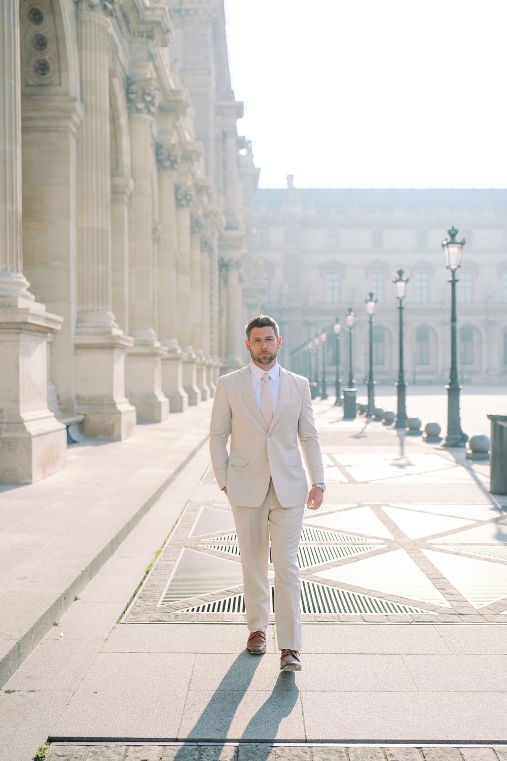 A well-dressed man in a beige suit walks confidently near the Louvre Museum in Paris on a sunny day.