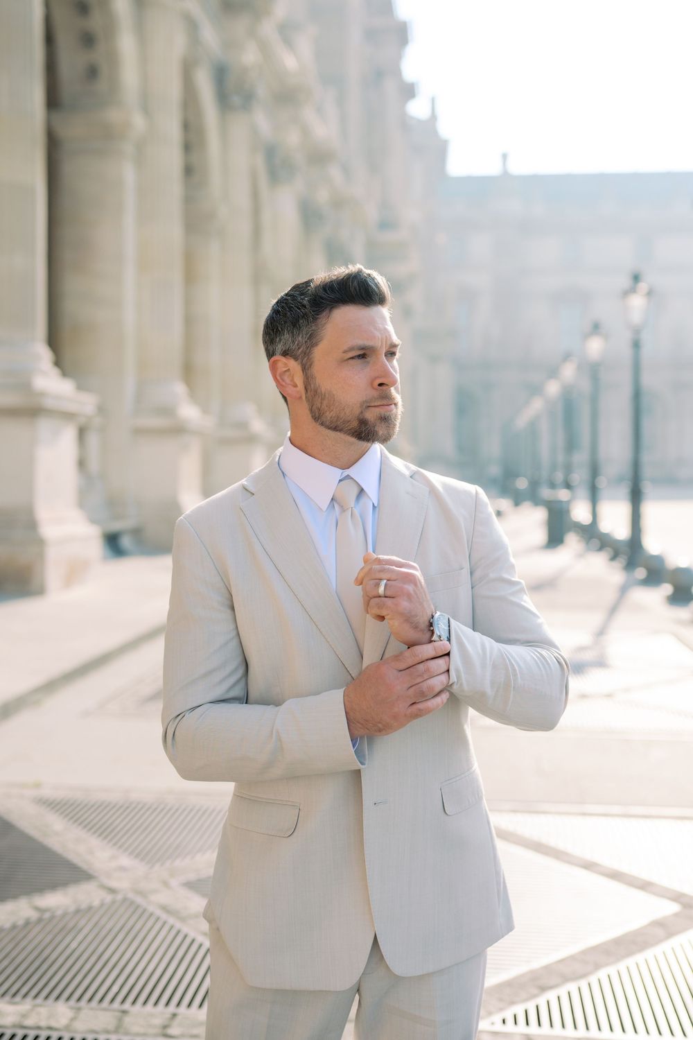 A well-dressed groom in a beige suit adjusts his cufflinks outside a grand Parisian building.