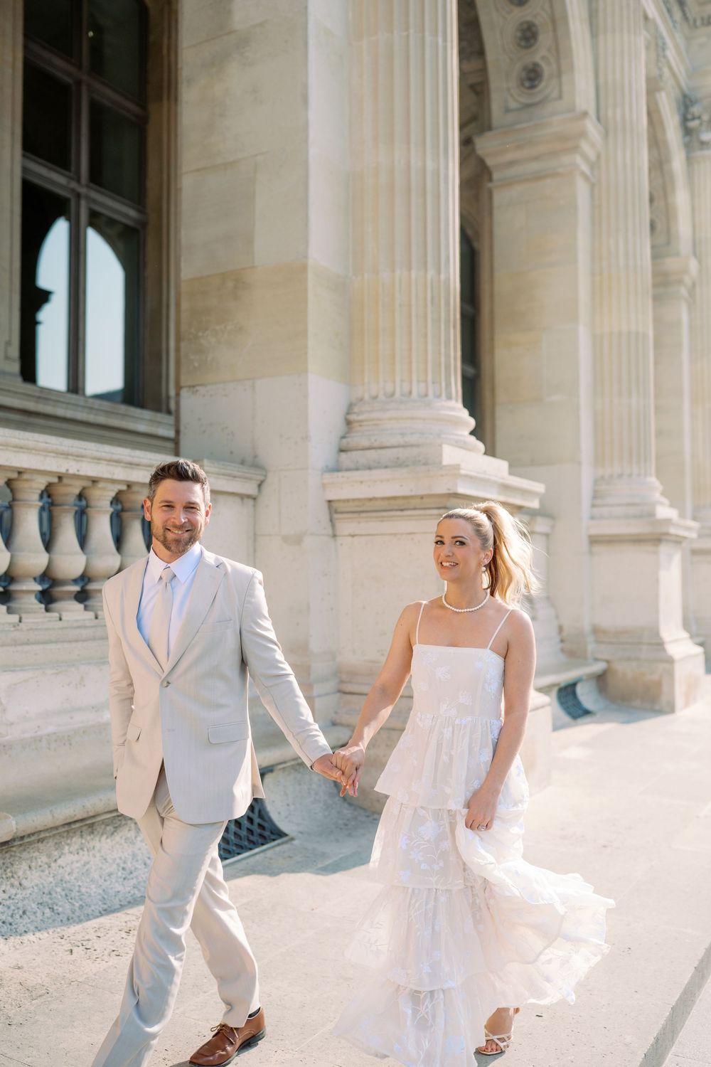 Couple in wedding attire holding hands walking beside a grand classical building in Paris.