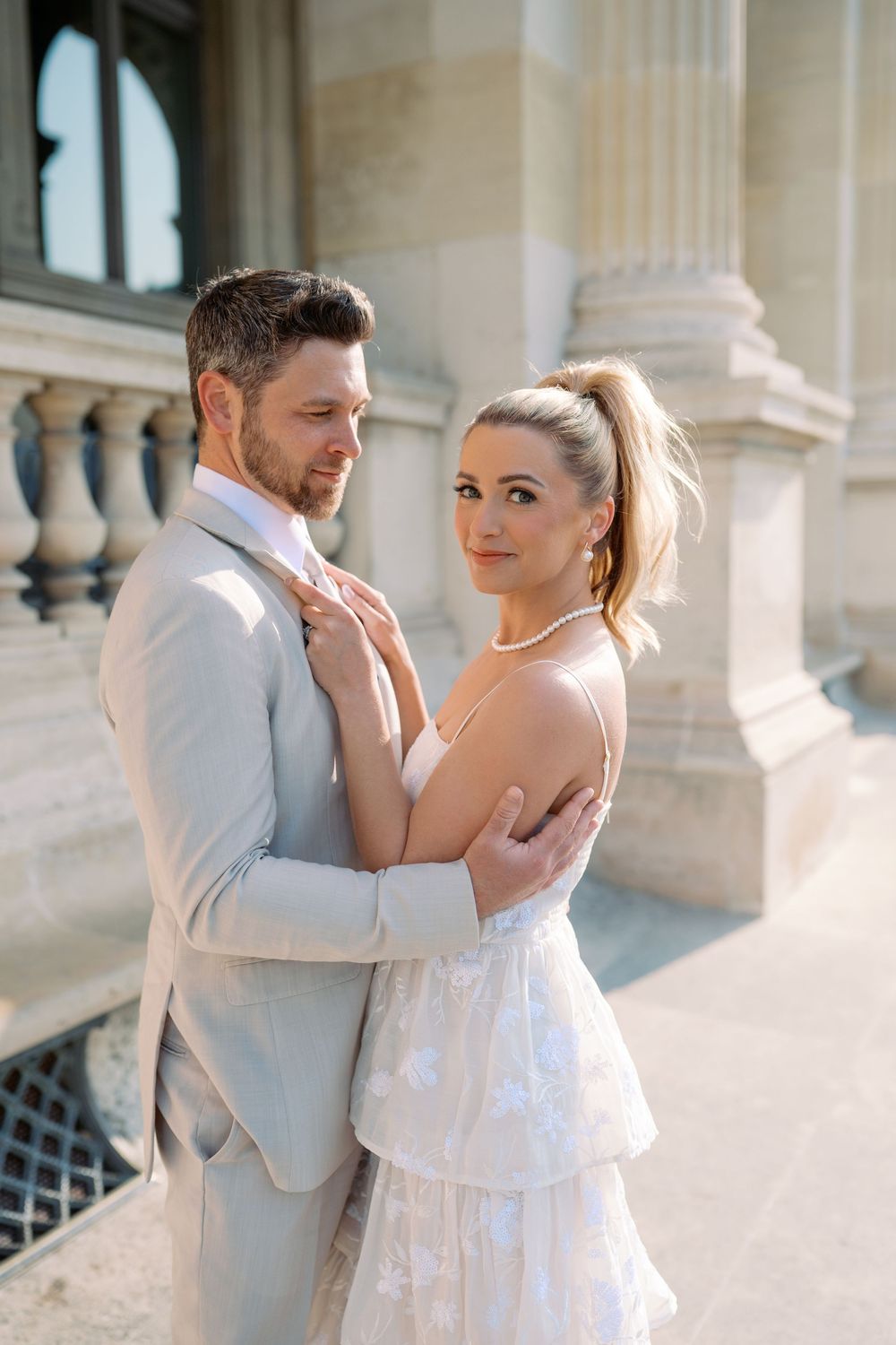 Elegant couple in light gray suit and floral dress embracing near classical stone columns in a romantic outdoor setting.