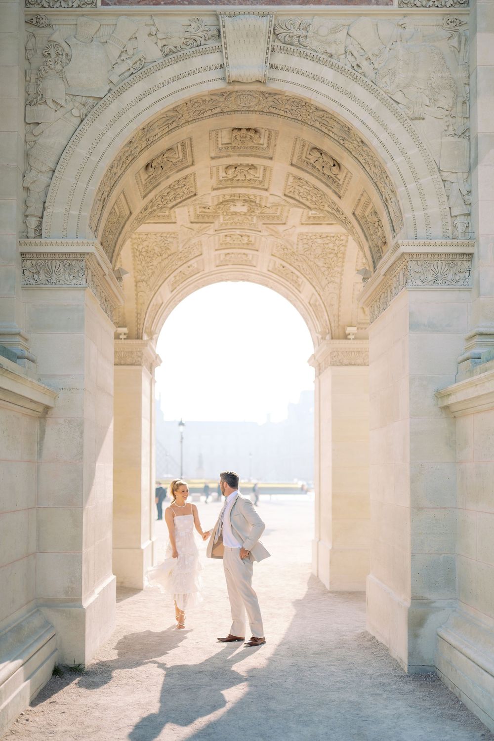 Bride and groom holding hands under ornate Parisian arch in soft golden light.