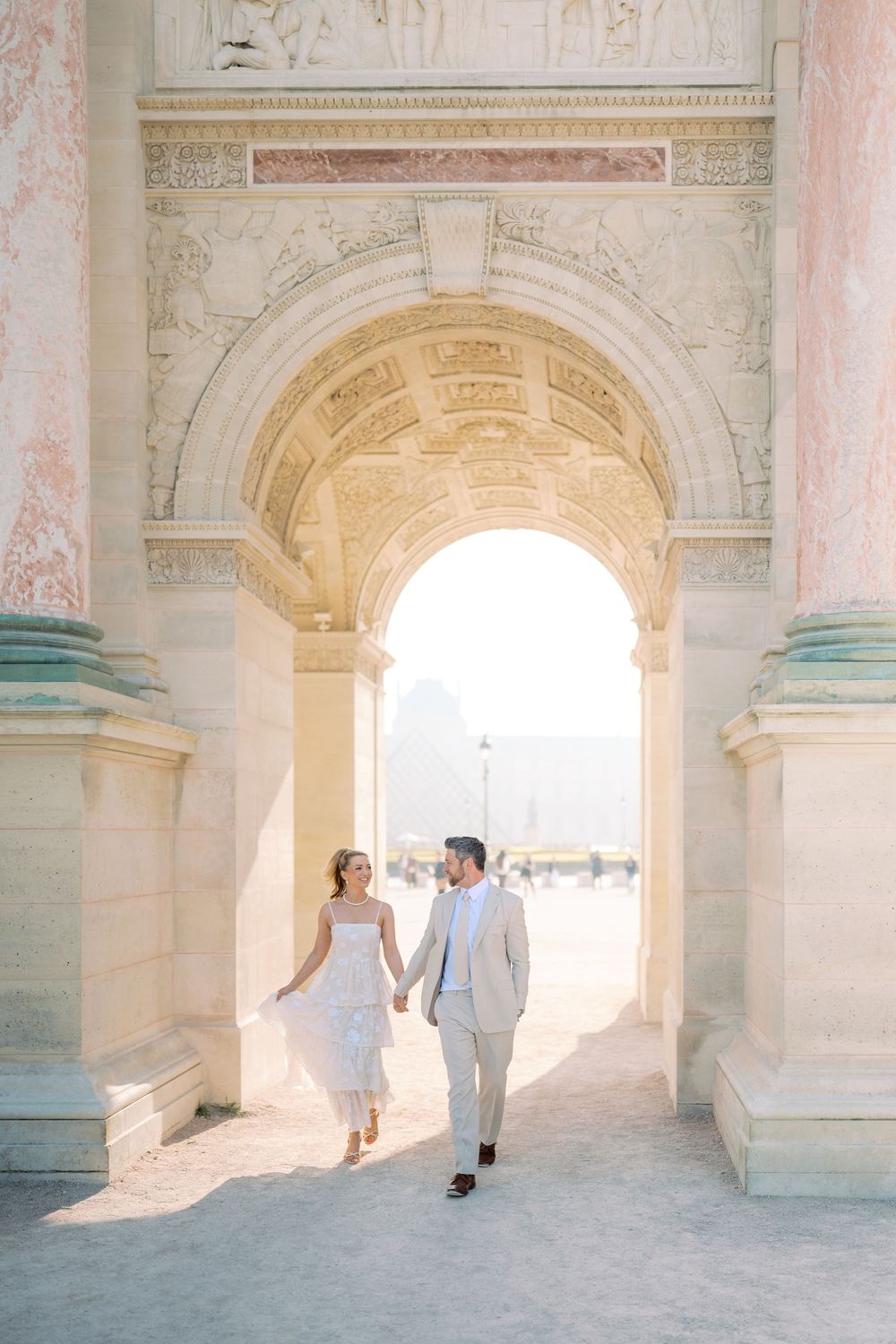 Newlyweds walking hand in hand through a sunlit grand arch at the Louvre in Paris.