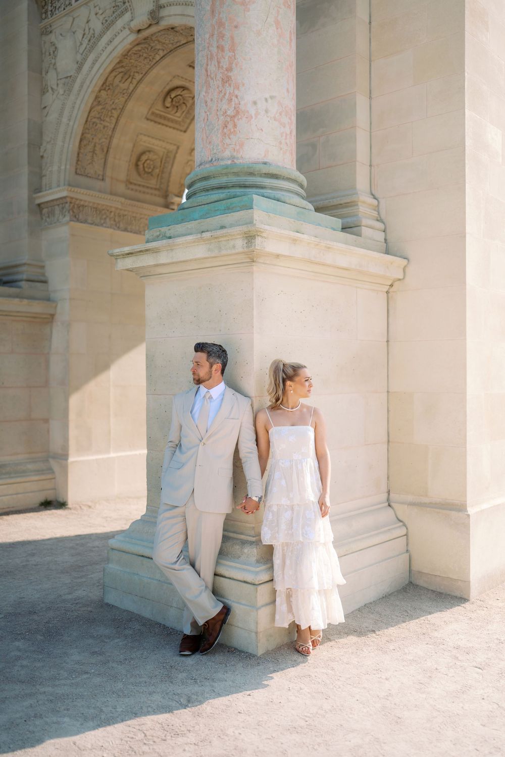 Bride and groom leaning against classical stone column in elegant wedding attire.