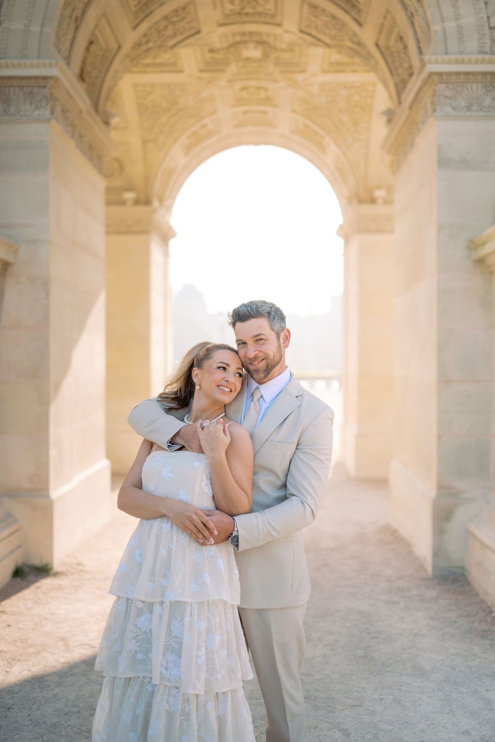 Couple embracing under ornate stone arch in Paris, woman in white tiered dress, man in beige suit, golden sunlight.