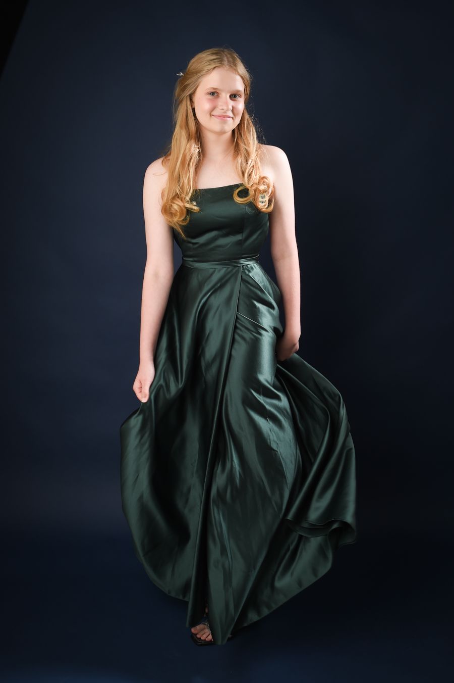 Young woman in a flowing emerald green prom gown stands against a dark backdrop. She smiles softly, exuding elegance and grace.