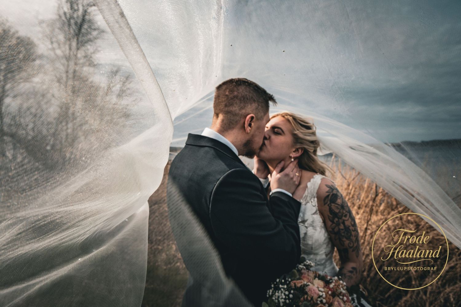 Couple sharing a passionate kiss beneath a billowing veil in a golden wheat field.