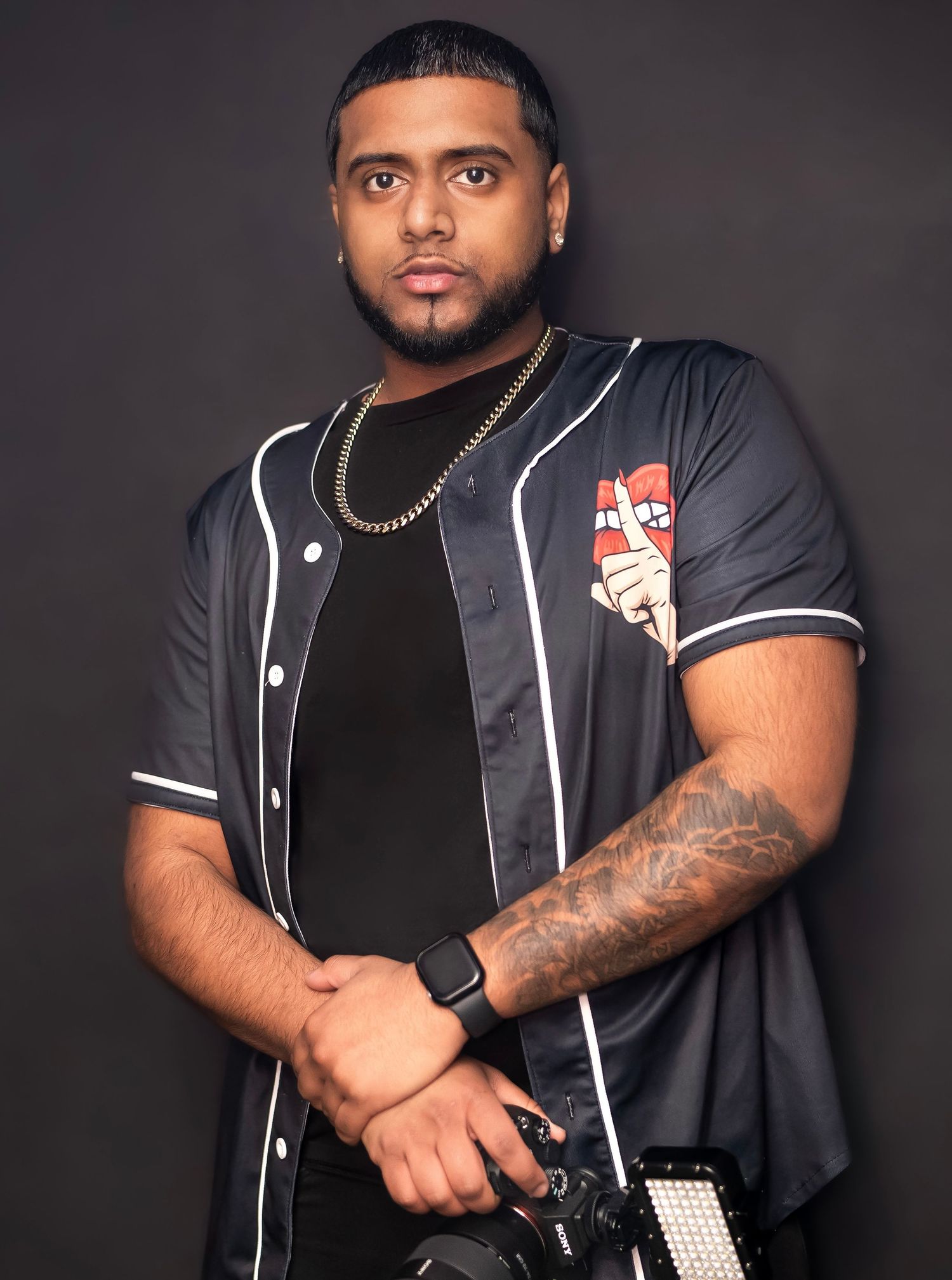 Photographer in baseball jersey holding camera with gold chain necklace posing against dark studio backdrop.
