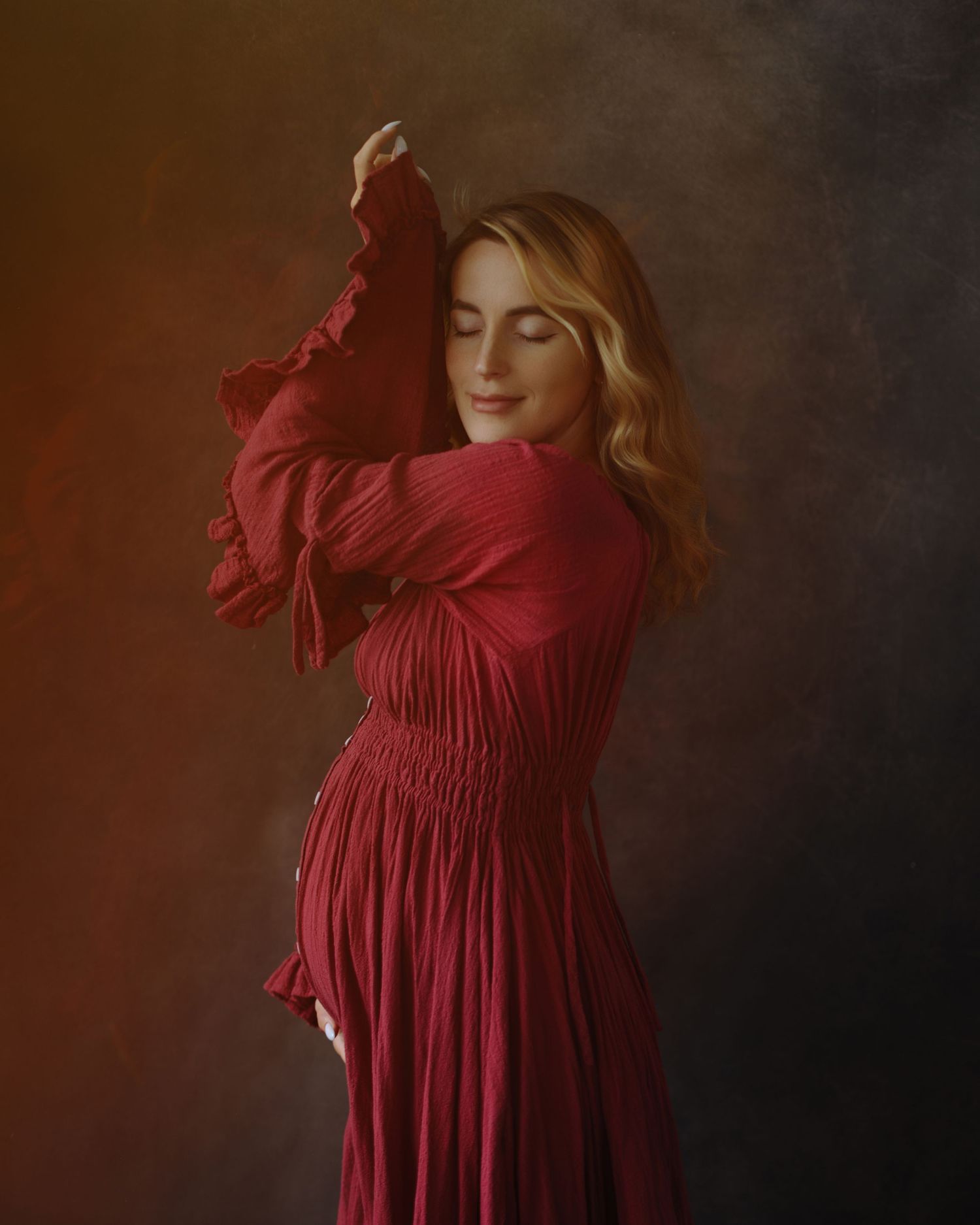 Pregnant woman with wavy blonde hair and closed eyes poses in a flowing crimson pleated dress against a moody painterly backdrop.