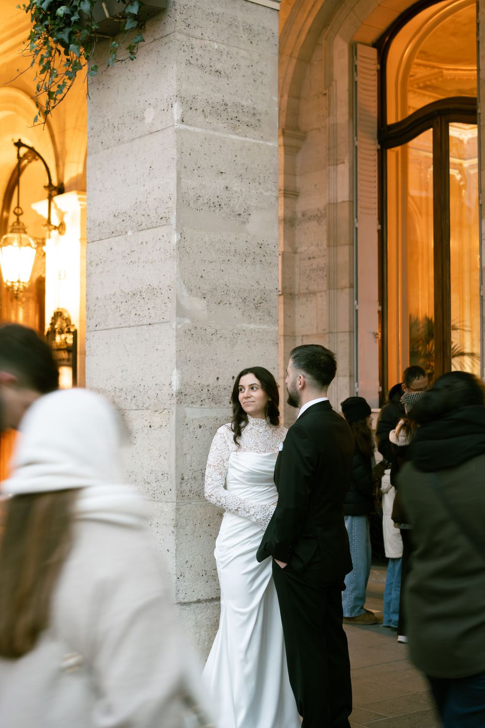 Bride in white lace gown and groom in black tuxedo pose by stone column at elegant Paris venue. Cinematic paris motion wedding photography