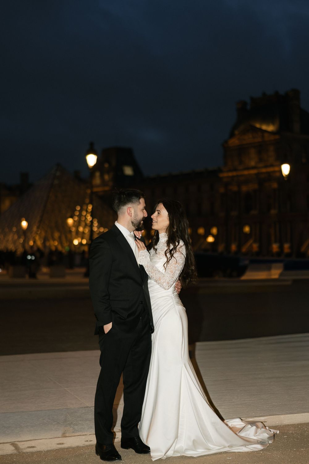 Bride and groom embracing at night in front of the illuminated Louvre pyramid in Paris. Editorial paris wedding photography, film
