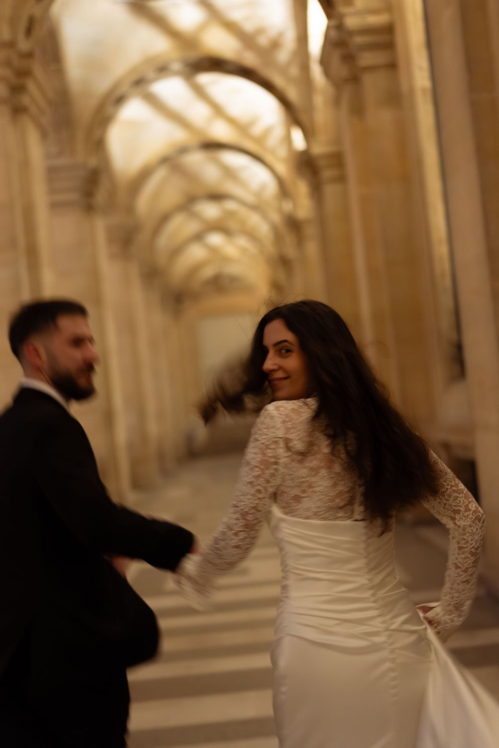 Bride in lace long-sleeve wedding dress smiling over shoulder while walking through ornate golden archway in Paris. Paris nighttime wedding
