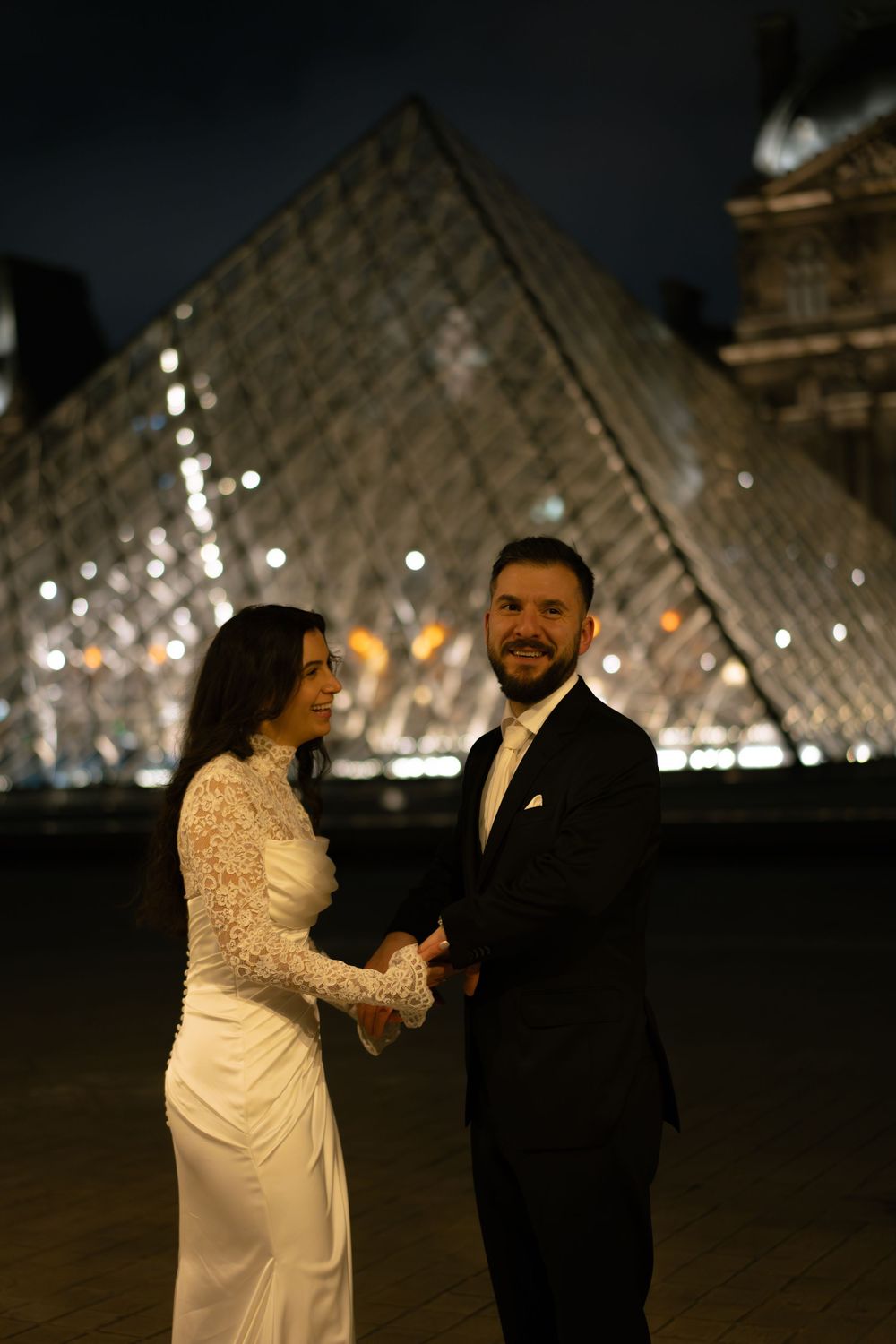 Bride and groom hold hands in front of illuminated Louvre Pyramid at night in Paris. Best paris editorial wedding photographer, film photos
