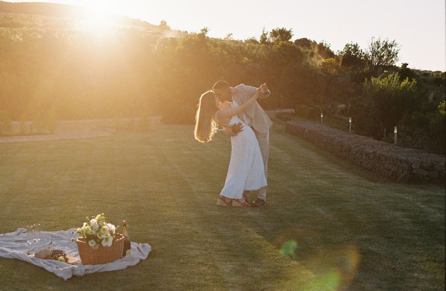 Bride and groom sharing a romantic kiss at golden hour on a lush green lawn with floral arrangement nearby.
