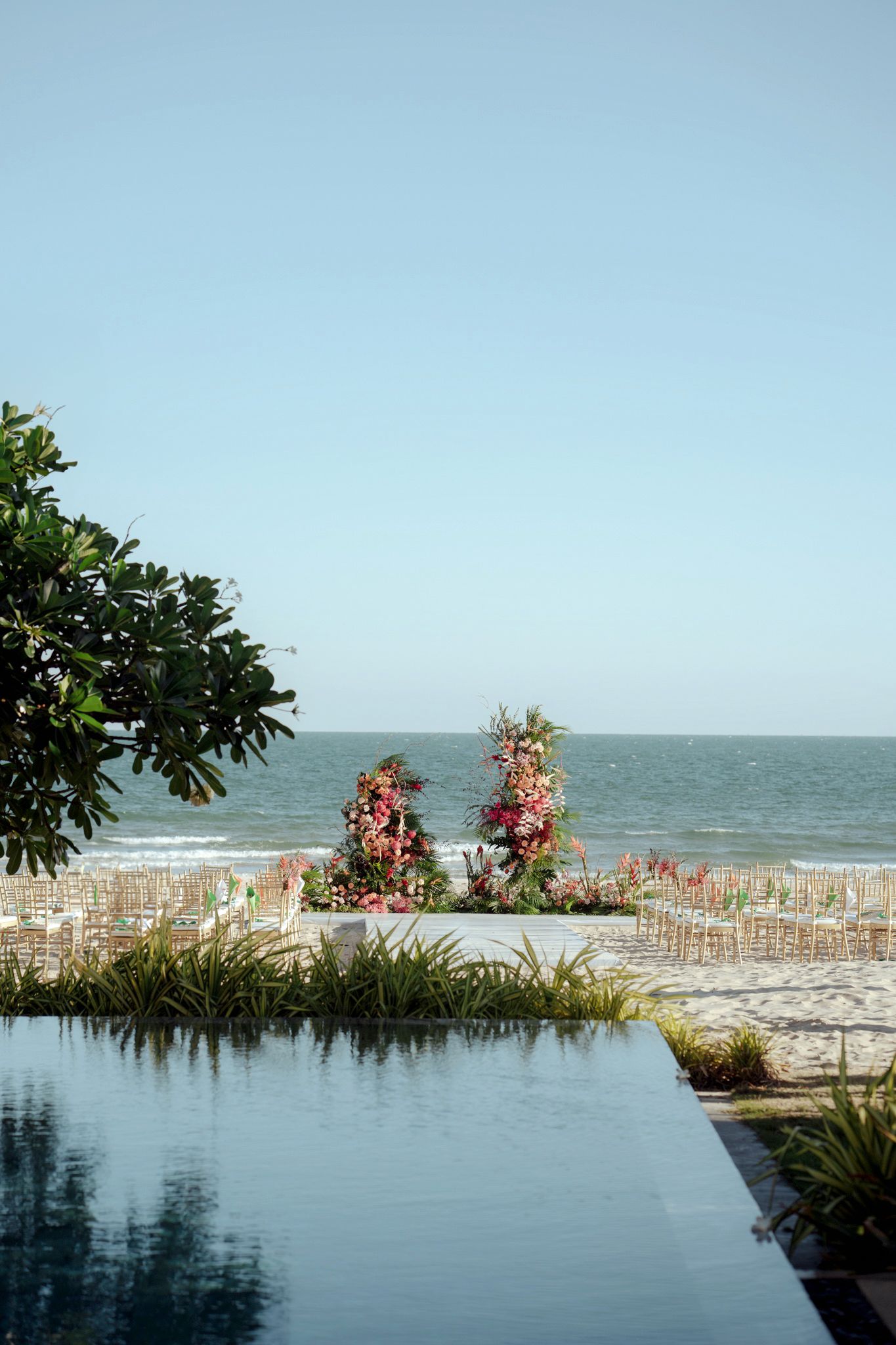 Beachfront wedding ceremony setup with floral arch, gold chairs, and ocean backdrop under clear blue sky.