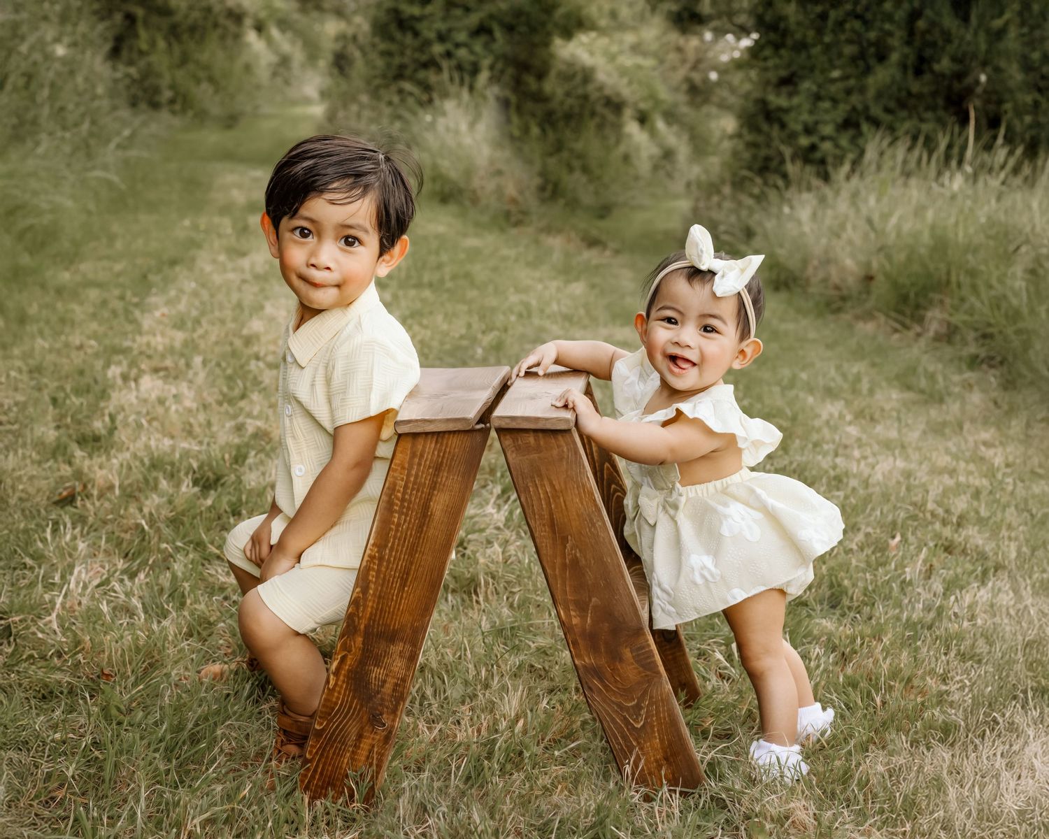 Two young children in matching cream outfits pose with a wooden bench in a lush green outdoor setting. Family Photographer Waterford