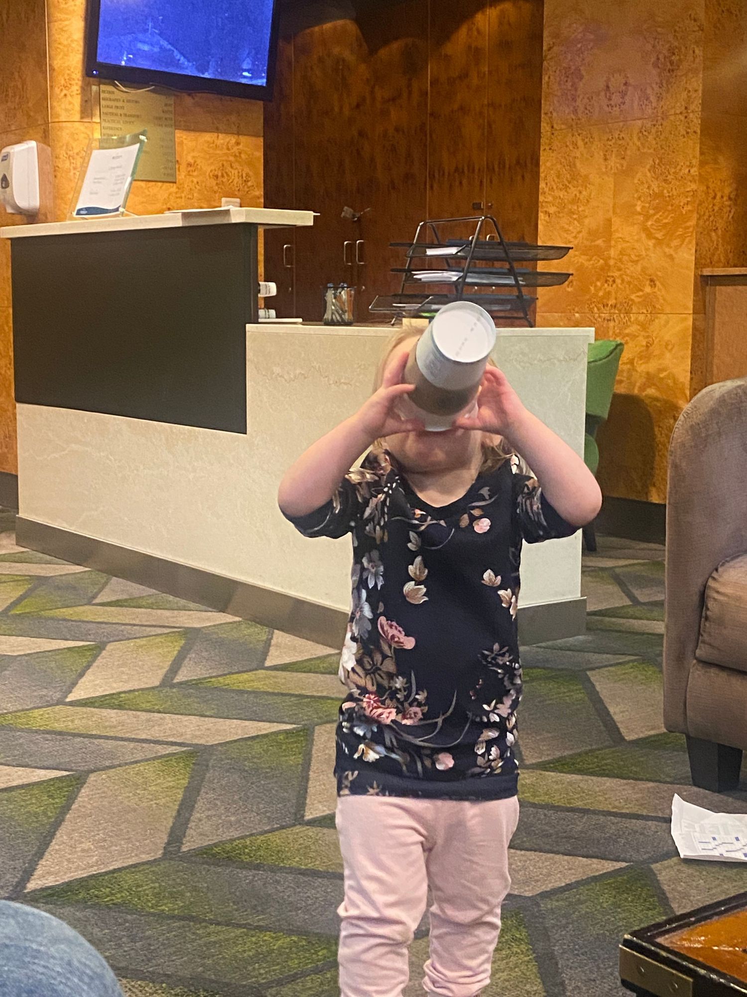 Young girl holds a paper cup up to her face like binoculars in a hotel lobby with patterned carpet.