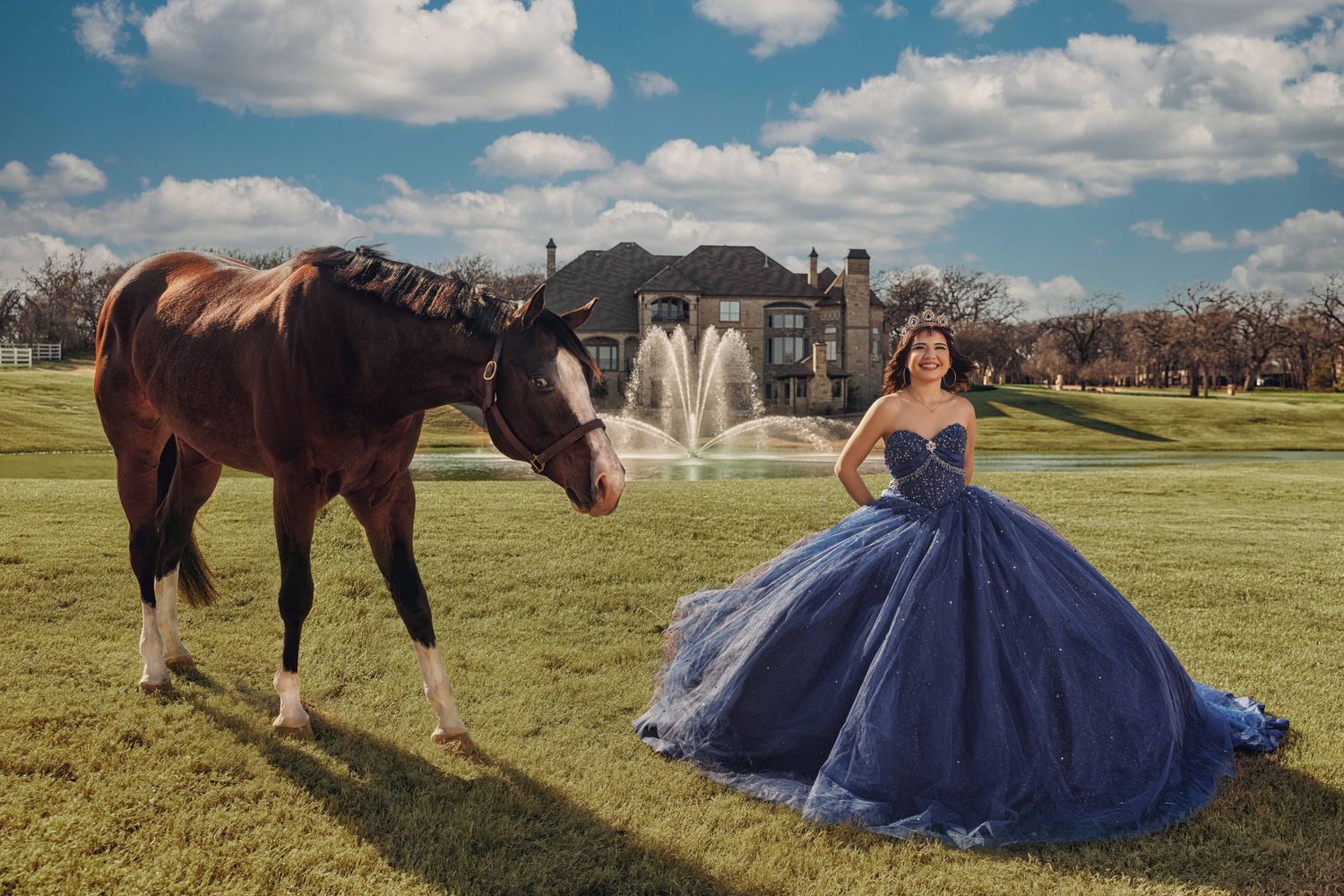 A girl in a navy blue beaded Quinceañera gown and tiara posing with a brown horse on a luxury estate lawn with a fountain background.