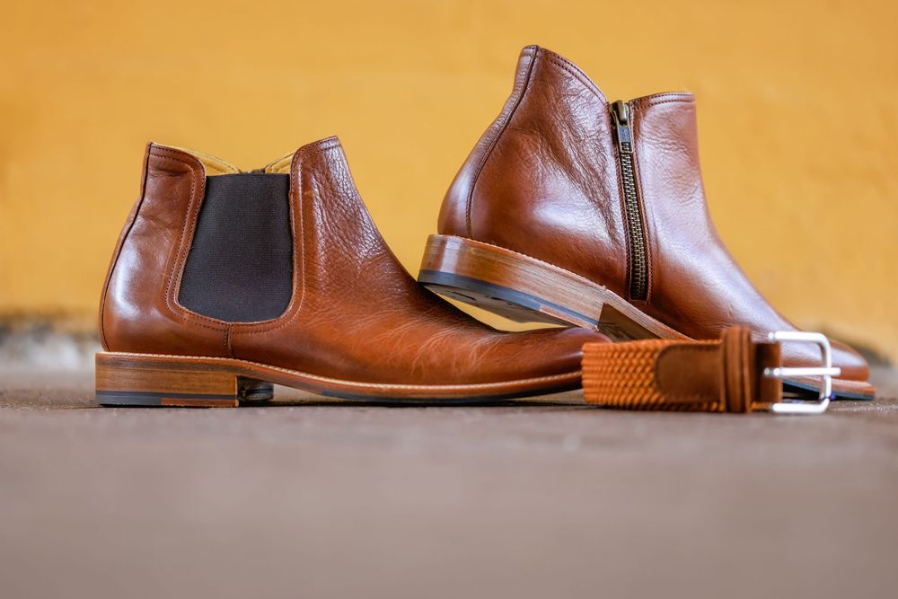 Brown leather Chelsea boots displayed with an orange braided belt on a gray surface against a yellow wall.