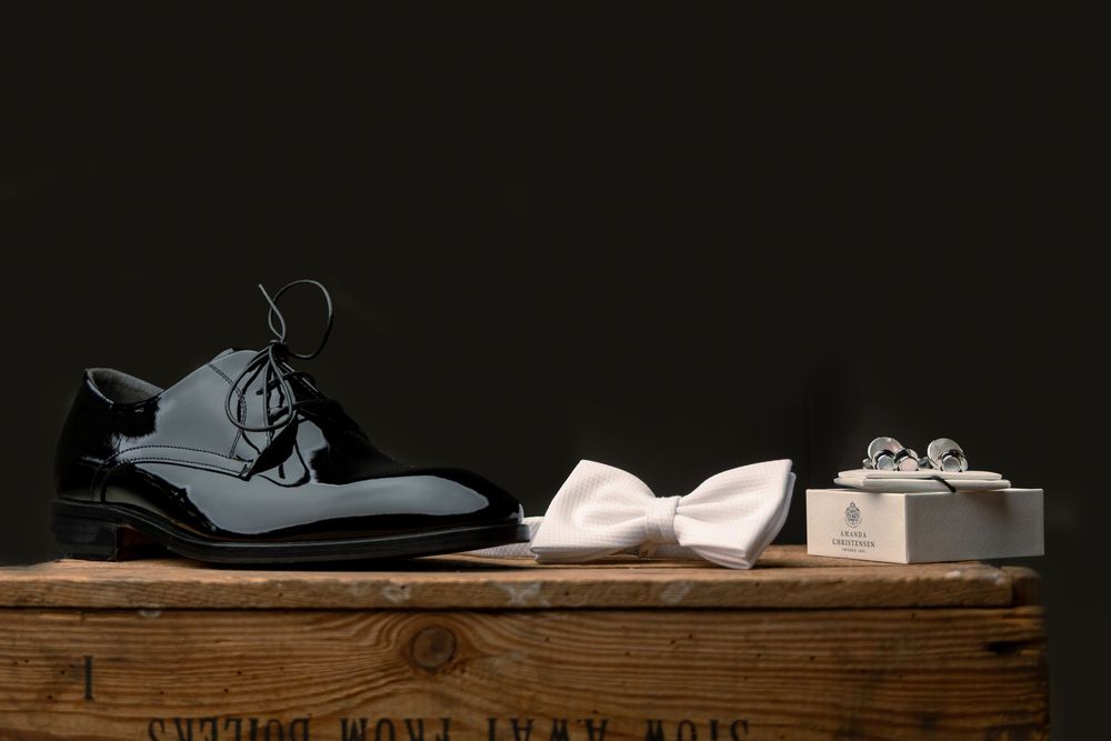 Black patent leather dress shoes beside a white bow tie and cufflinks on a wooden box, elegant groom accessories.