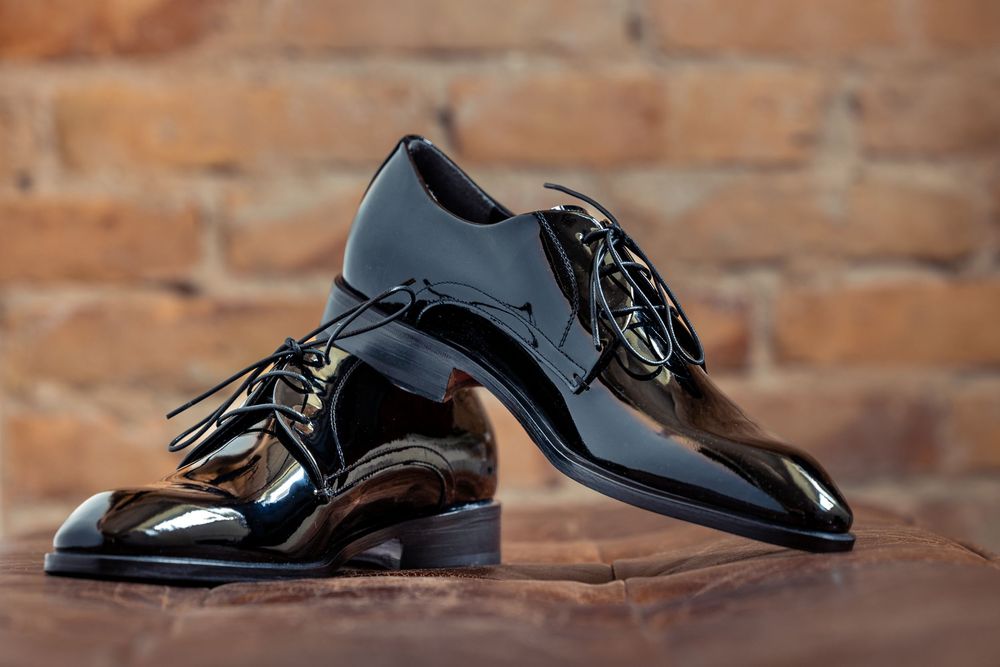 Shiny black patent leather Oxford dress shoes with laces displayed on wooden surface against brick wall.