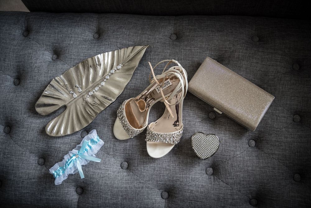 Bridal accessories arranged on gray tufted sofa including heels, clutch, garter, and silver accents.