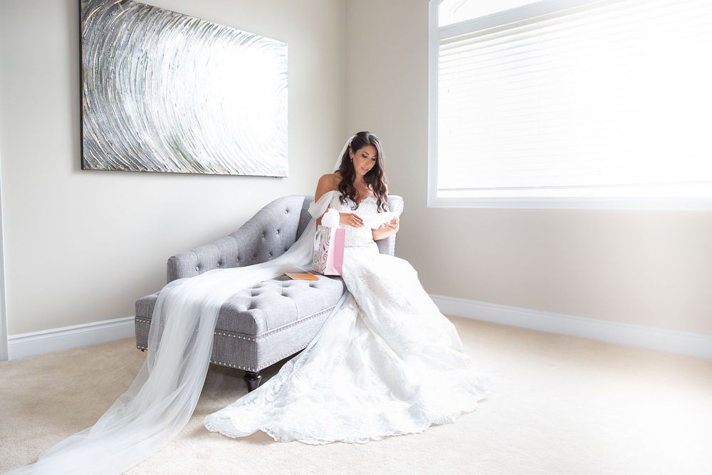 Bride in full lace gown sits on gray chaise opening a pink gift box.