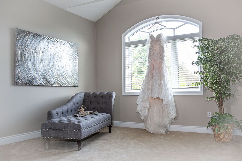 Lace wedding gown hangs in sunlit window of elegant room with gray chaise and artwork.