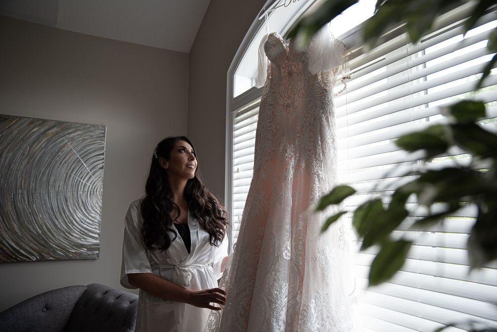 Bride in white robe gazes at her lace wedding gown hanging in a sunlit window.