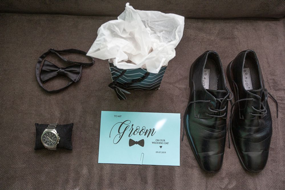 Groom wedding accessories flat lay featuring black bow tie, dress shoes, watch, and a teal Groom card on a sofa.