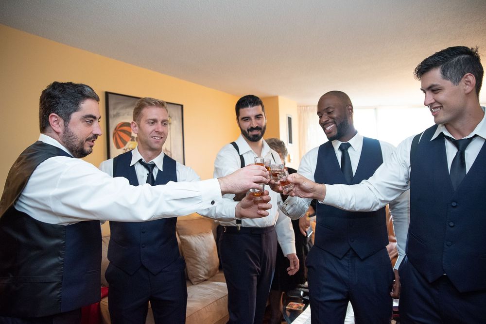 Groom and groomsmen in navy suits toasting together in a hotel room during wedding day preparations.