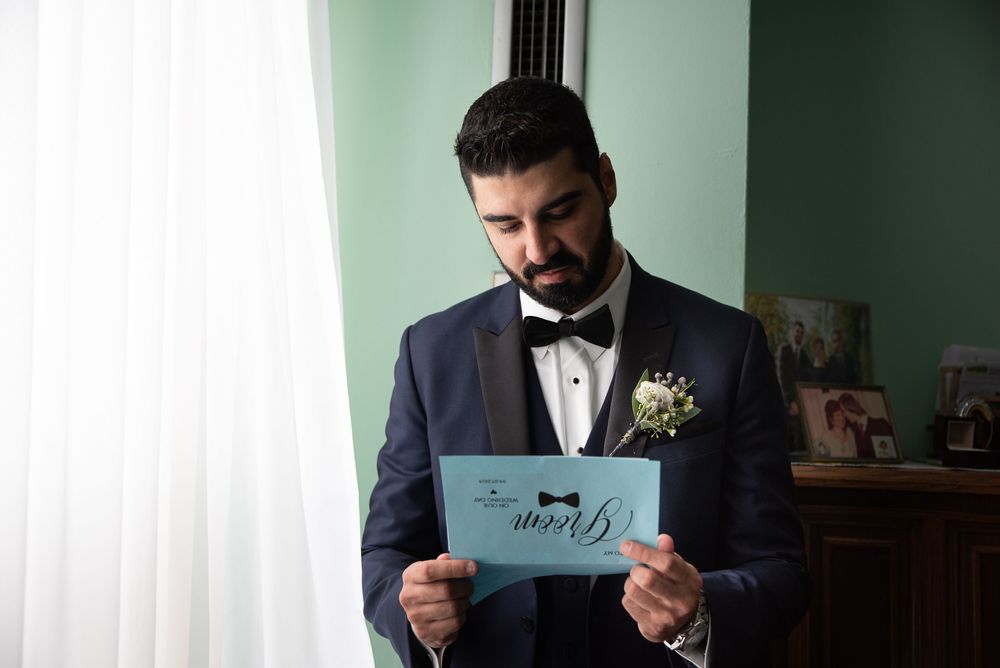 Dapper groom in a navy tuxedo reading a teal Groom card while standing by a window before his wedding.