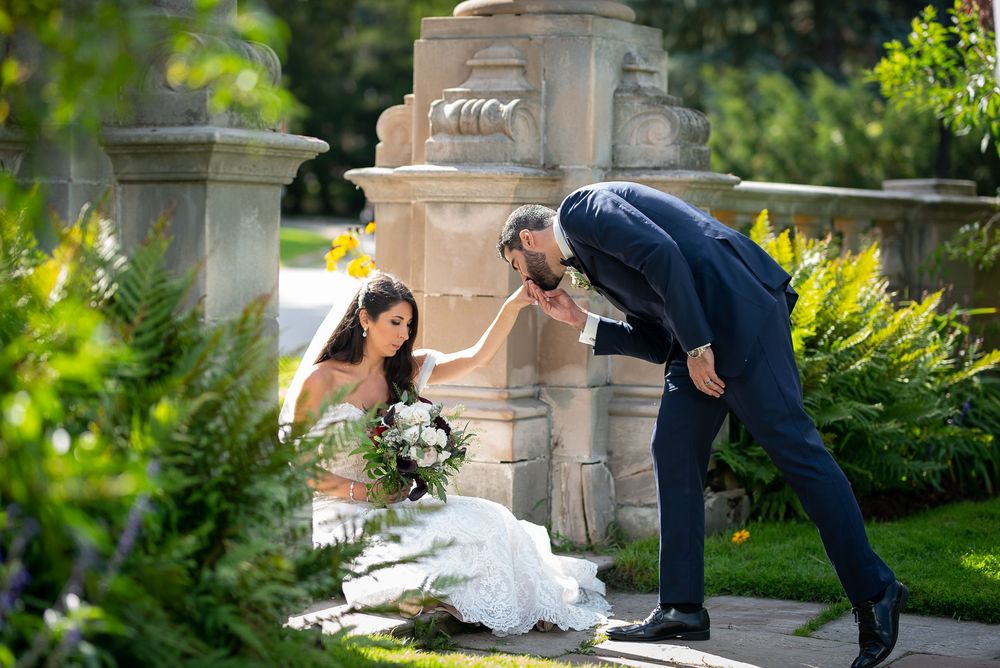 Groom in navy suit leans down to kiss bride's hand as she sits in lace gown by stone pillar.