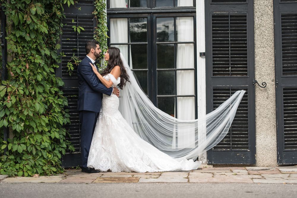 Bride in lace gown with flowing veil and groom in navy suit pose by ivy-covered shutters.