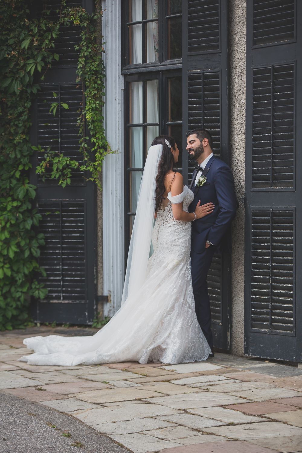 Bride in lace gown with cathedral veil and groom in navy suit pose by black shuttered doors.