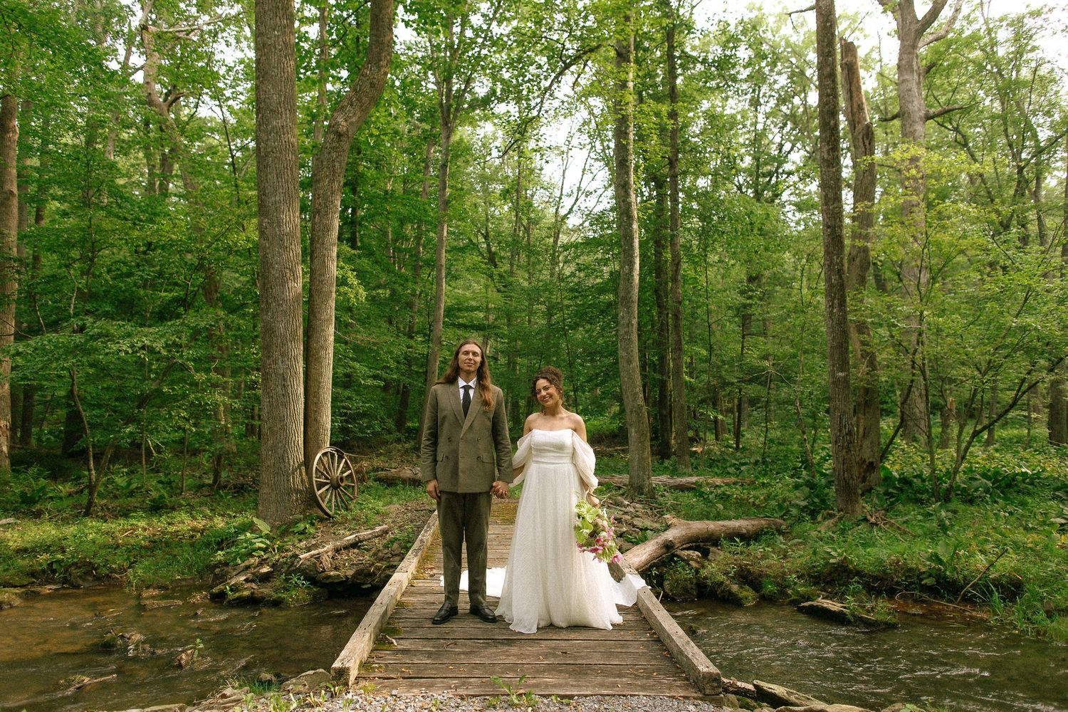 Bride and groom standing on a wooden bridge in a lush green forest during their outdoor woodland wedding.