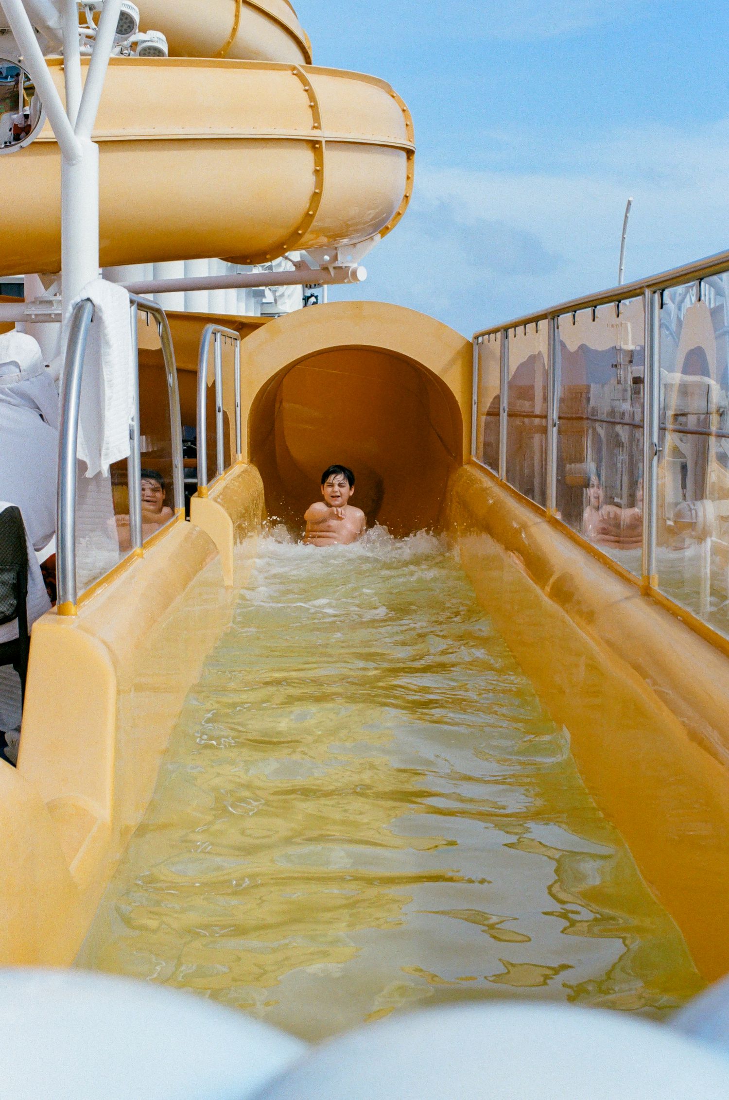 Child sliding down a yellow water slide on a cruise ship deck under a blue sky.