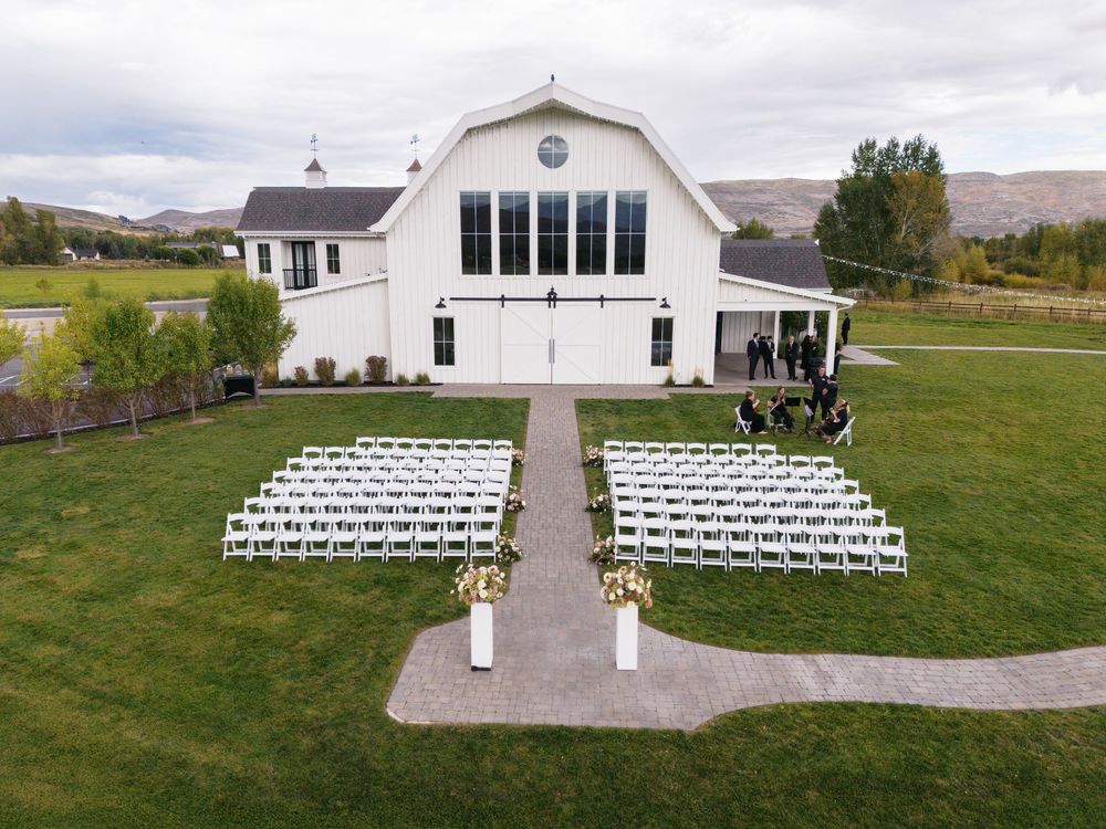 Aerial view of white barn wedding venue with rows of white chairs set up for outdoor ceremony on lush green lawn.