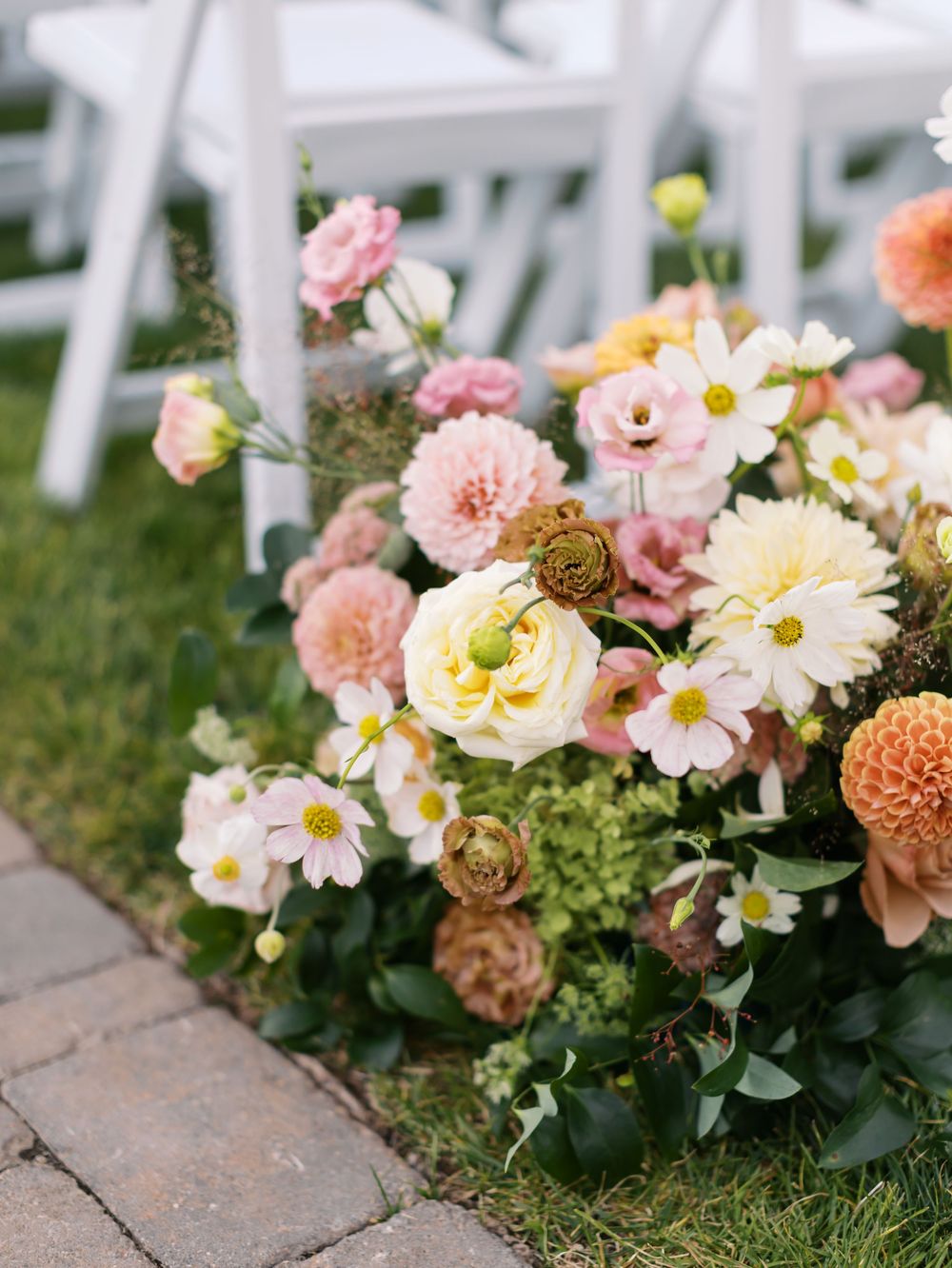 Lush outdoor wedding floral arrangement featuring pink dahlias, white daisies, and peach blooms on a stone path.