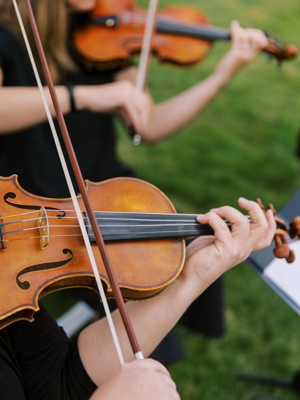 Two violinists in black attire perform outdoors on a green lawn, showcasing close-up detail of violin bows and strings.