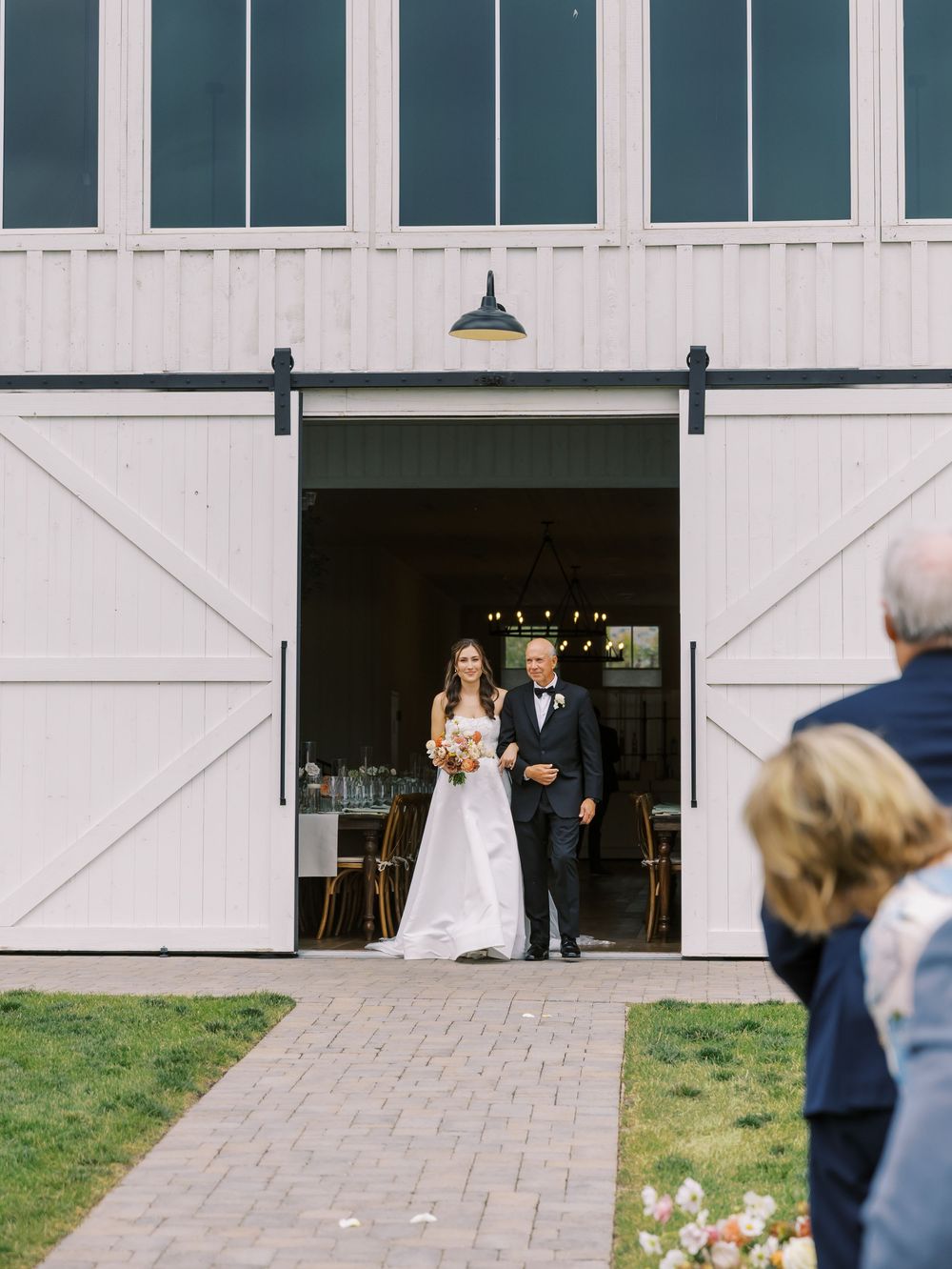 Bride and father walk through white barn doors at an outdoor wedding ceremony on a sunny day.