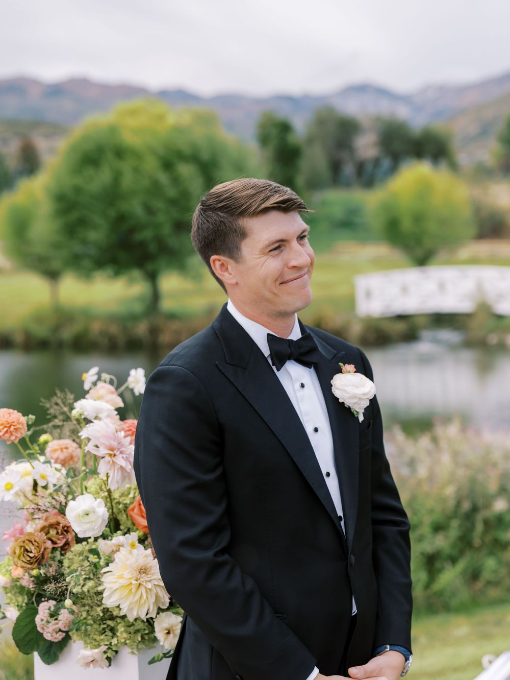 Groom in black tuxedo smiling at altar with floral arrangements and mountain lake backdrop.