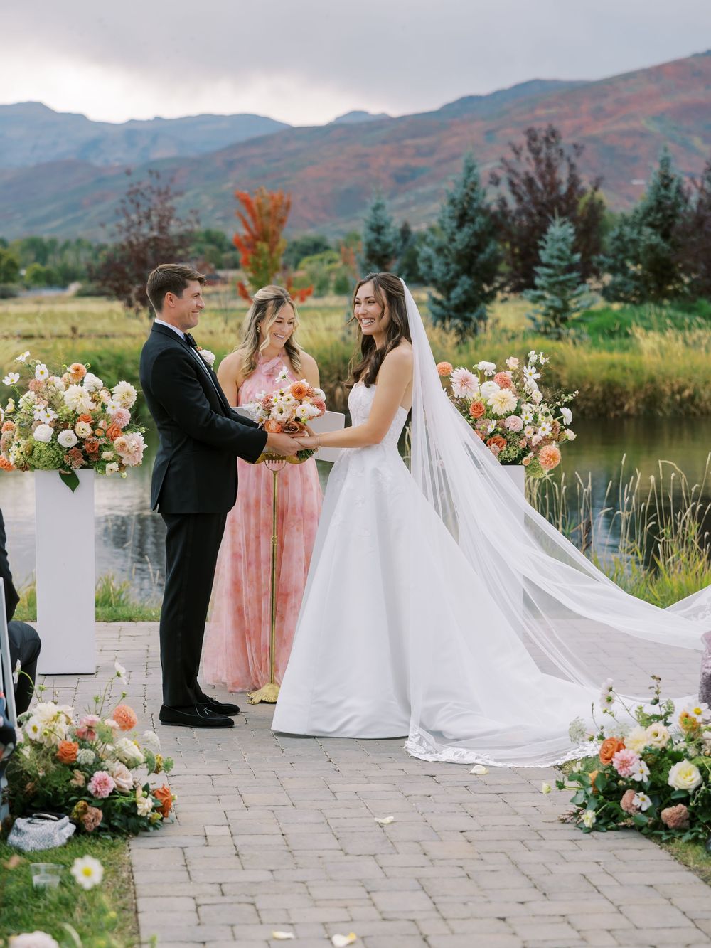 Bride and groom exchange vows outdoors with mountain backdrop, floral arrangements, and a bridesmaid nearby.