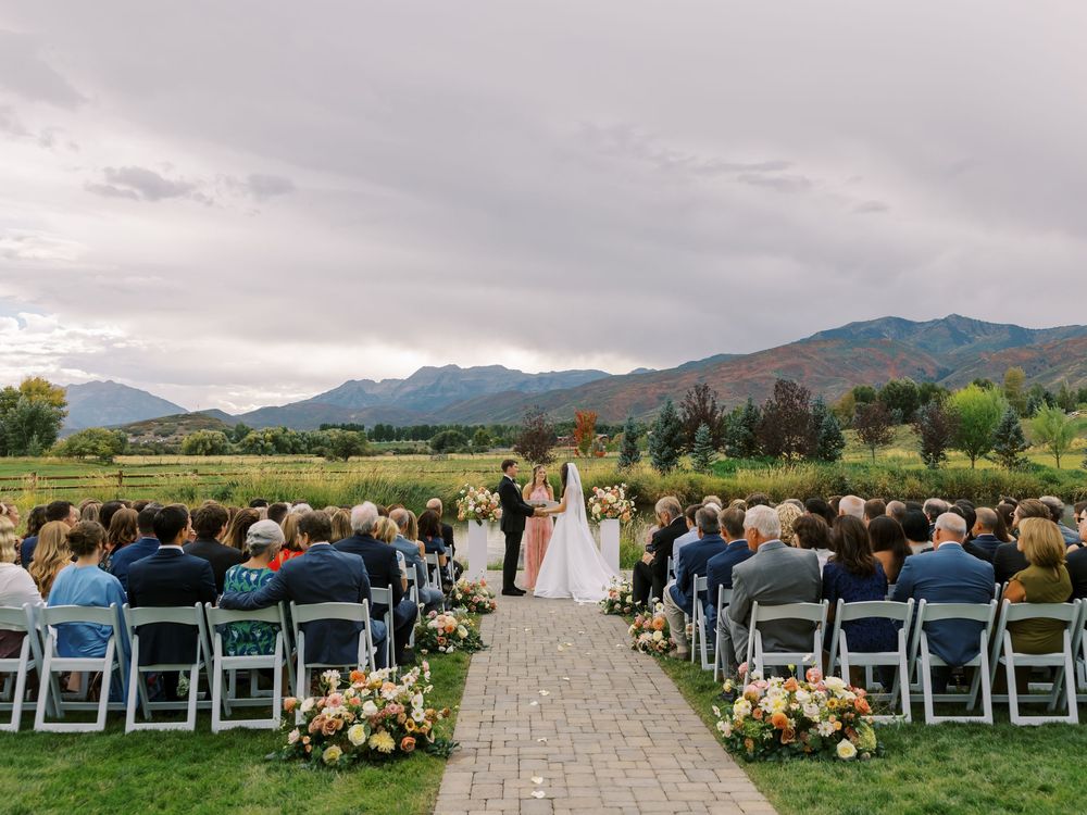 Guests seated at an outdoor wedding ceremony with mountain views, floral aisle decor, and overcast skies.