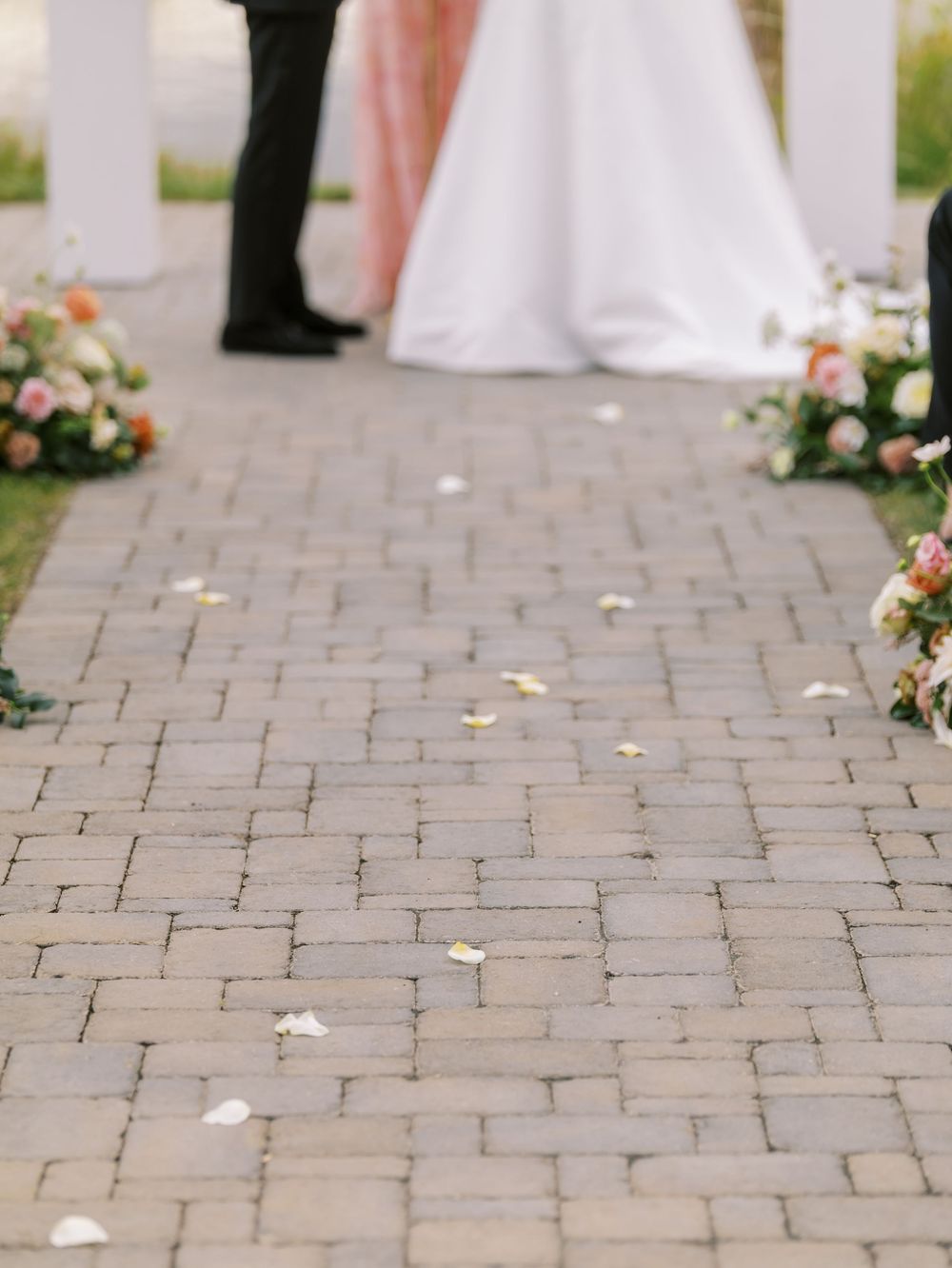 Flower petal-lined brick aisle leads to couple at outdoor wedding ceremony altar with floral arrangements.